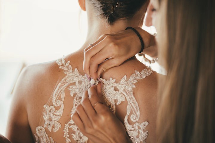 A bride with tanned skin being buttoned into a lace gown
